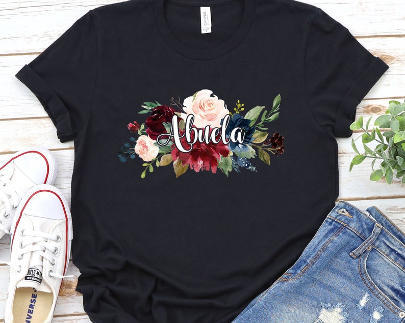 Pu&ograve; includere: Maglietta nera con un design floreale e la parola "Abuela" in corsivo bianco.