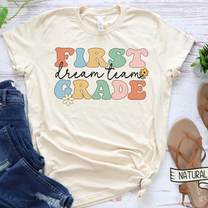 Op de afbeelding: Een beige T-shirt met de tekst "FIRST dream team GRADE" in kleurrijke retro-lettertype met bloem accenten.