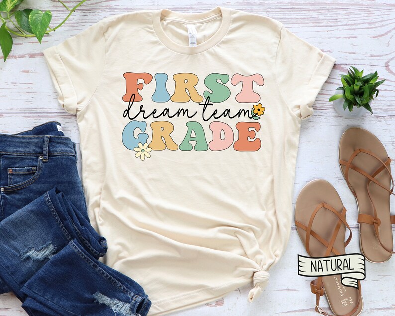 Op de afbeelding: Een beige T-shirt met de tekst "FIRST dream team GRADE" in kleurrijke retro-lettertype met bloem accenten.