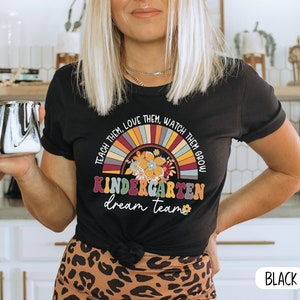 May include: Black t-shirt with a rainbow and floral design. The text "TEACH THEM. LOVE THEM. WATCH THEM GROW KINDERGARTEN dream team" is printed on the shirt.
