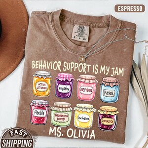 May include: A brown t-shirt with the text "Behavior Support is My Jam" and a row of eight jars with different labels, including "Active Listening", "Empathy", "Inspiring", "Patient", "Flexible", "Personalized Support", "Inclusion", and "Compassion". The text "MS. Olivia" is below the jars.