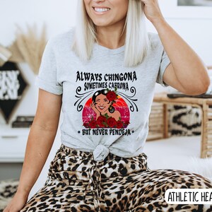 May include: A gray t-shirt with a graphic of a woman with flowers in her hair. The text on the shirt reads "Always Chingona Sometimes Cabrona - But Never Pendeja".