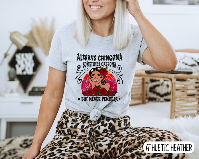May include: A gray t-shirt with a graphic of a woman with flowers in her hair. The text on the shirt reads "Always Chingona Sometimes Cabrona - But Never Pendeja".