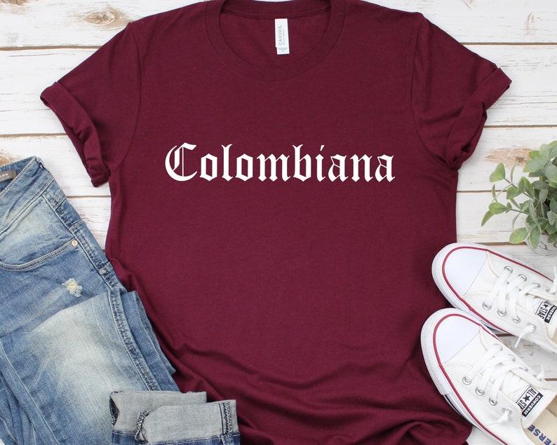 Op de afbeelding: Een maroon t-shirt met een witte afbeelding waarop in gotische letters "Colombiana" staat.