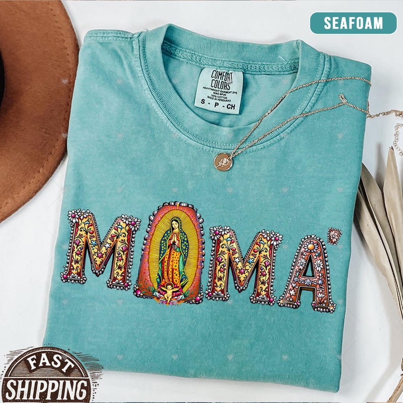 May include: Seafoam green t-shirt with a floral design spelling "MOMA" with a depiction of the Virgin Mary in the center. The shirt has a tag that reads "Comfort Colors" and "S-P-CH".