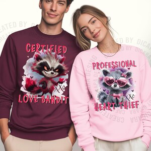 May include: Two sweatshirts, one burgundy and one pink, each featuring a raccoon graphic. The burgundy sweatshirt says "CERTIFIED LOVE BANDIT" and the pink one says "PROFESSIONAL HEART THIEF".