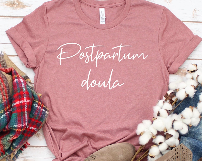 May include: A dusty rose colored t-shirt with the text "Postpartum Doula" written in white cursive lettering.