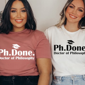 May include: Two people wearing t-shirts with the text "Ph.Done. Doctor of Philosophy" and a graduation cap graphic. The shirt on the left is pink and the shirt on the right is white.