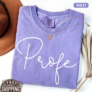 May include: A violet Comfort Colors t-shirt with the word "Profe" in white script. A gold necklace with a pendant rests on the shirt. The shirt is folded, and a brown hat is partially visible in the background.