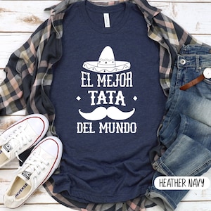 May include: A navy blue t-shirt with white text that reads "El Mejor Tata Del Mundo" with a white sombrero and mustache graphic. The shirt is laid flat on a wooden surface with a plaid shirt, jeans, and white sneakers.