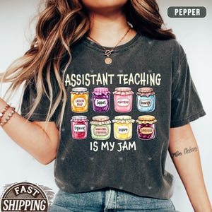 Puede incluir: Una camiseta gris oscuro con el texto "Assistant Teaching is my jam" y un gr&aacute;fico de tarros coloridos con palabras como "Preparaci&oacute;n de la lecci&oacute;n", "Apoyo", "Monitoreo", "Animar", "Organizar", "Estrategias conductuales", "Inspirar" y "Supervisar".