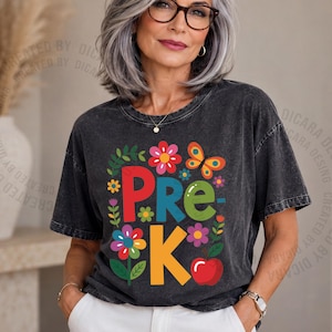 May include: A charcoal grey acid-wash t-shirt featuring a colorful graphic with the text PRE-K in large letters, surrounded by illustrations of various flowers, a yellow butterfly, and a red apple, all arranged in a rectangular composition.