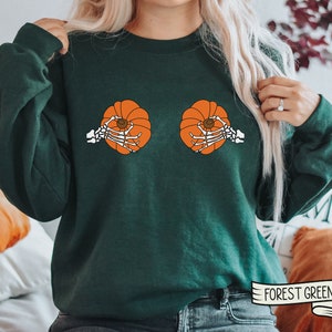 Op de afbeelding: Groen sweatshirt met een afbeelding van twee pompoenen met skeletten die ze vasthouden. De tekst "FOREST GREEN" staat op een wit lint in de rechterbenedenhoek van de afbeelding.