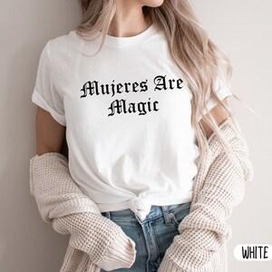 May include: White t-shirt with the text "Mujeres Are Magic" printed in black.