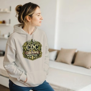 May include: A light beige hoodie with a front pocket and a hood. The front of the hoodie features a decorative emblem with the text "CDC CORKTOWN DRINKING CLUB" in a vintage style, with a skull and other decorative elements.