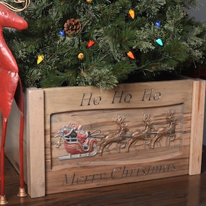 Puede incluir: Un soporte de árbol de Navidad de madera con las palabras "Ho Ho Ho" y "Merry Christmas" grabadas en la parte delantera. El soporte presenta un relieve del trineo de Papá Noel y renos. Dos renos de metal rojo flanquean el árbol.