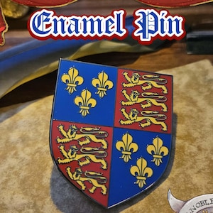 May include: Enamel pin featuring a shield design with a blue and red background. The shield is divided into four sections with gold fleur-de-lis and lions. The text "Enamel Pin" is at the top of the image.