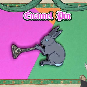 May include: Enamel pin featuring a gray rabbit playing a silver horn against a pink and green background. The pin has the text "Enamel Pin" in a pink and white font. The pin is set in a decorative frame.