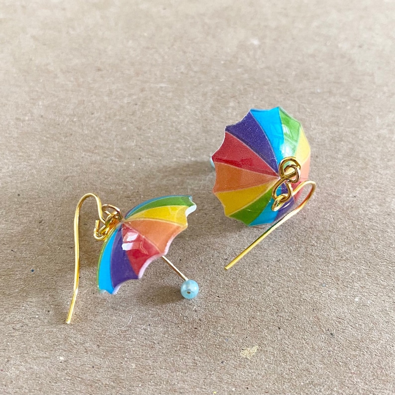 Puede incluir: Un par de pendientes de paraguas de colores arco&iacute;ris con ganchos dorados. Los paraguas est&aacute;n hechos de resina y tienen un acabado brillante.