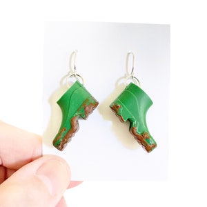 Può includere: Orecchini a forma di stivali verdi e marroni con ganci d'argento. Gli stivali sono realizzati in pasta polimerica e hanno una texture fangosa.