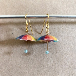 Puede incluir: Un par de pendientes de paraguas de colores arco&iacute;ris con ganchos dorados y cuentas de color azul claro. Los pendientes cuelgan de una barra plateada.