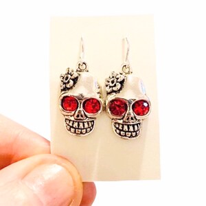 May include: Silver skull earrings with red rhinestone eyes and a small flower detail.