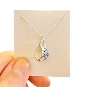 Op de afbeelding: Een delicate porseleinen theekopje hanger ketting. De miniatuur beker heeft een witte basis met blauwe bloempatronen en gouden randen. De hanger hangt aan een fijne zilveren ketting, wat een elegant en charmant accessoire creëert.