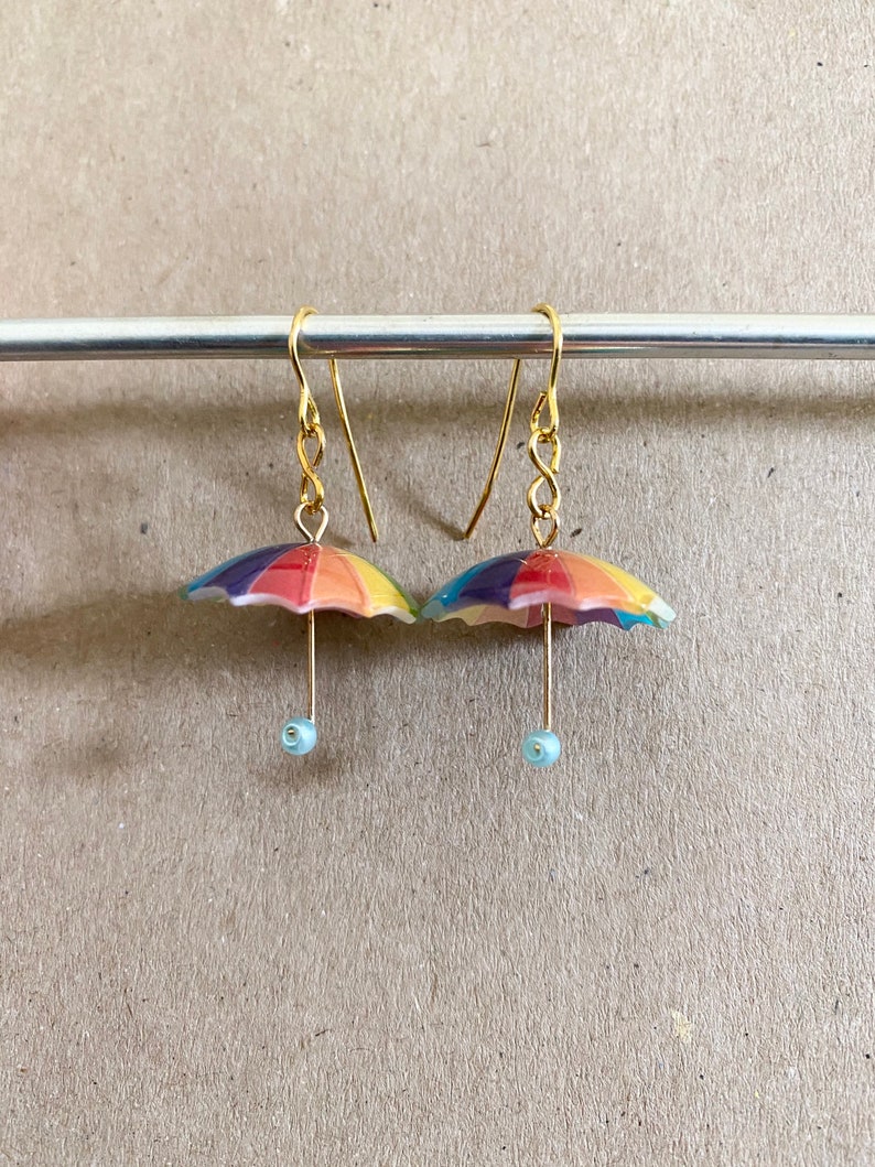 Puede incluir: Un par de pendientes de paraguas de colores arco&iacute;ris con ganchos dorados y cuentas de color azul claro. Los pendientes cuelgan de una barra plateada.