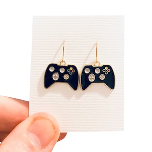 Op de afbeelding: Een paar gouden oorbellen met zwarte emaille-videogamecontroller-bedels. De controllers hebben witte en gekleurde knoppen.