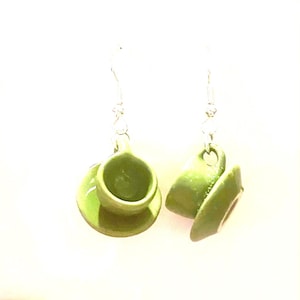 Op de afbeelding: Een paar groene keramische oorringen in de vorm van een theekopje. Elke oorring heeft een miniatuur theekopje en schotel ontwerp, opgehangen aan een zilverkleurige haak. De kopjes zijn glanzend groen en de schotels zijn rond.