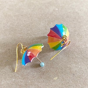 Puede incluir: Un par de pendientes de paraguas de colores arco&iacute;ris con ganchos dorados. Los paraguas est&aacute;n hechos de resina y tienen un acabado brillante.