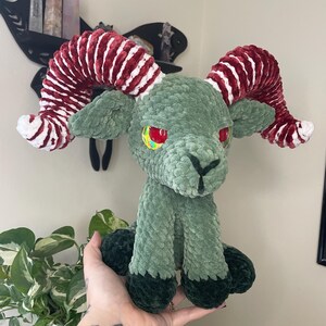 May include: A handmade plush ram with a green body, dark green hooves, and red and white striped horns. The ram has red and yellow eyes and is sitting upright. The plush is made of soft, textured material.