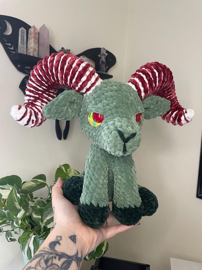 May include: A handmade plush ram with a green body, dark green hooves, and red and white striped horns. The ram has red and yellow eyes and is sitting upright. The plush is made of soft, textured material.
