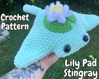 May include: A crocheted stingray in a light blue color, with a lily pad and flower detail on top. The stingray has large, black, reflective eyes. The text "Crochet Pattern" and "Lily Pad Stingray" are visible.