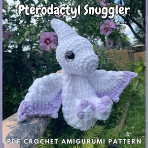 May include: A white crocheted amigurumi pattern of a Pterodactyl with purple wings. The pattern is called "Pterodactyl Smuggler" and is a PDF crochet amigurumi pattern.