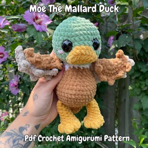Può includere: Un'anatra amigurumi all'uncinetto con testa verde, corpo marrone e zampe gialle. L'anatra ha occhi neri e un becco giallo. Il testo "Moe The Mallard Duck" e "Pdf Crochet Amigurumi Pattern" sono visibili.