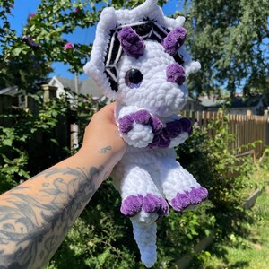 May include: White crocheted dinosaur with purple accents and a black and white spiderweb design on its head.