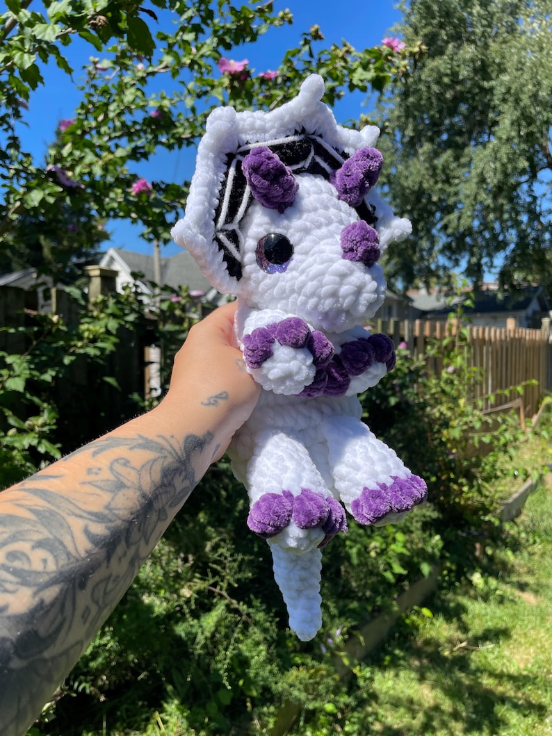 May include: White crocheted dinosaur with purple accents and a black and white spiderweb design on its head.