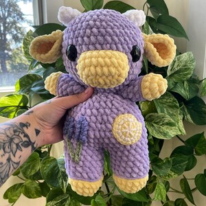 Puede incluir: Un juguete de peluche de vaca de crochet morado con orejas y cuernos amarillos. La vaca tiene una nariz amarilla y ojos negros. La vaca est&aacute; sosteniendo una flor de lavanda y un lim&oacute;n.