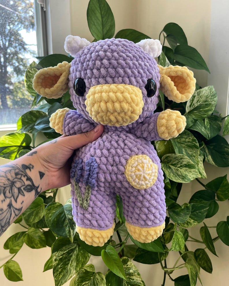 Puede incluir: Un juguete de peluche de vaca de crochet morado con orejas y cuernos amarillos. La vaca tiene una nariz amarilla y ojos negros. La vaca est&aacute; sosteniendo una flor de lavanda y un lim&oacute;n.