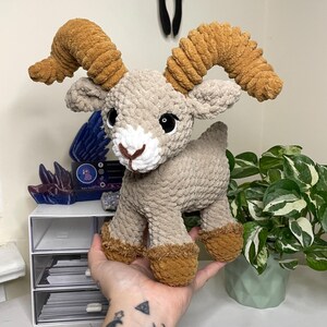 May include: A handmade plush ram toy with a light tan body, brown hooves, and large, textured brown horns. The ram has black eyes and a white muzzle. The toy is made of soft, textured yarn and is designed to be a cuddly stuffed animal.
