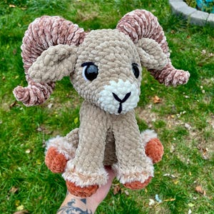 May include: A plush, crocheted ram with large, curled horns. The ram is primarily tan with brown horns and accents of white and orange. It has large black eyes and is sitting on a grassy surface.