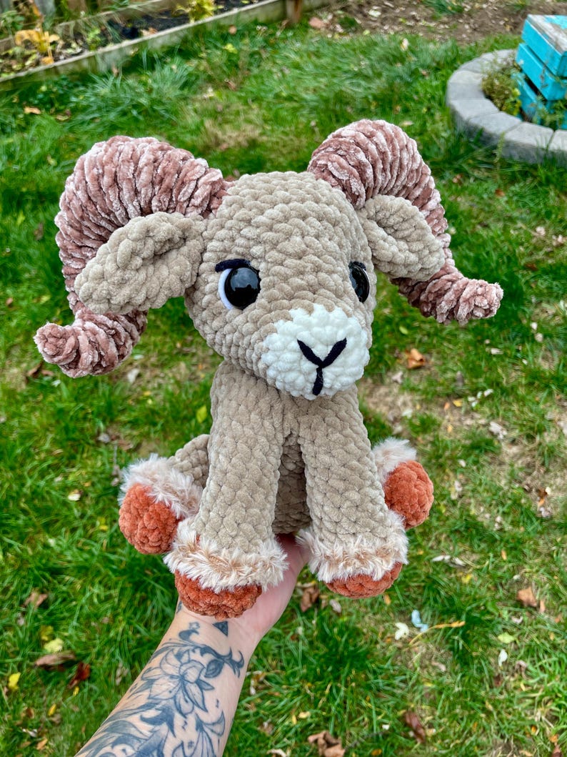May include: A plush, crocheted ram with large, curled horns. The ram is primarily tan with brown horns and accents of white and orange. It has large black eyes and is sitting on a grassy surface.