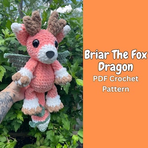 May include: A handmade crochet fox-dragon plushie in shades of pink, white, brown, and green. The plushie has large black eyes, antlers, and wings. The text "Briar The Fox Dragon PDF Crochet Pattern" is displayed on an orange background.