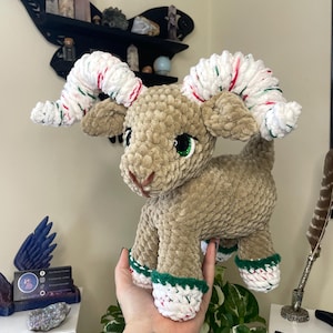 May include: A plush, tan-colored ram with white and red-striped horns and green-rimmed hooves. The ram has large, green eyes and is made of a soft, textured material. The ram is a handmade item.
