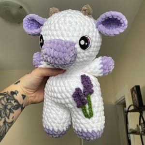 Puede incluir: Un juguete de peluche blanco de crochet con forma de vaca con orejas, pezu&ntilde;as y nariz moradas. El juguete tiene flores de lavanda bordadas en un lado y ojos brillantes.