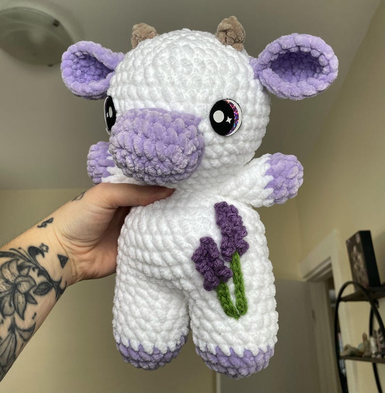 Puede incluir: Un juguete de peluche blanco de crochet con forma de vaca con orejas, pezu&ntilde;as y nariz moradas. El juguete tiene flores de lavanda bordadas en un lado y ojos brillantes.