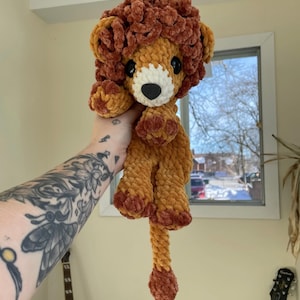 May include: A crocheted lion plush toy in brown and orange tones. The lion has a fluffy mane and a long tail. The text "2in1 No Sew Lion Pdf Crochet Pattern" is visible at the top of the image.