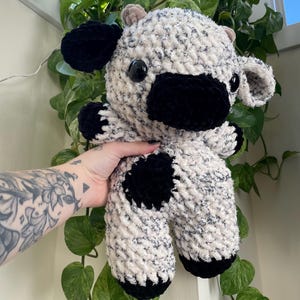 Puede incluir: Una vaca de peluche tejida a crochet con un cuerpo blanco y negro manchado, orejas negras y pezu&ntilde;as negras.