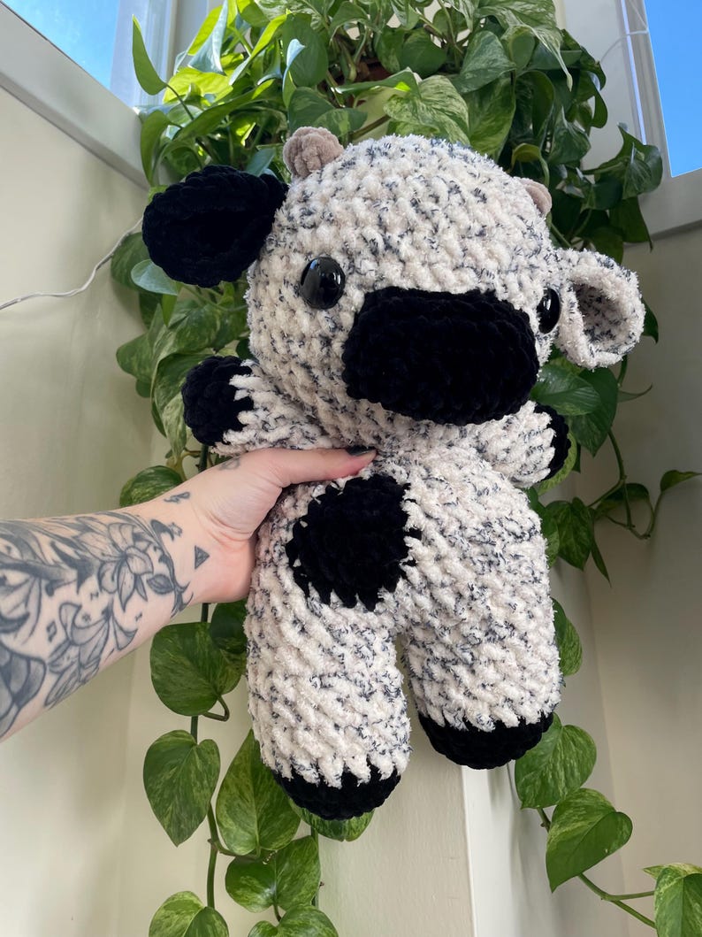 Puede incluir: Una vaca de peluche tejida a crochet con un cuerpo blanco y negro manchado, orejas negras y pezu&ntilde;as negras.